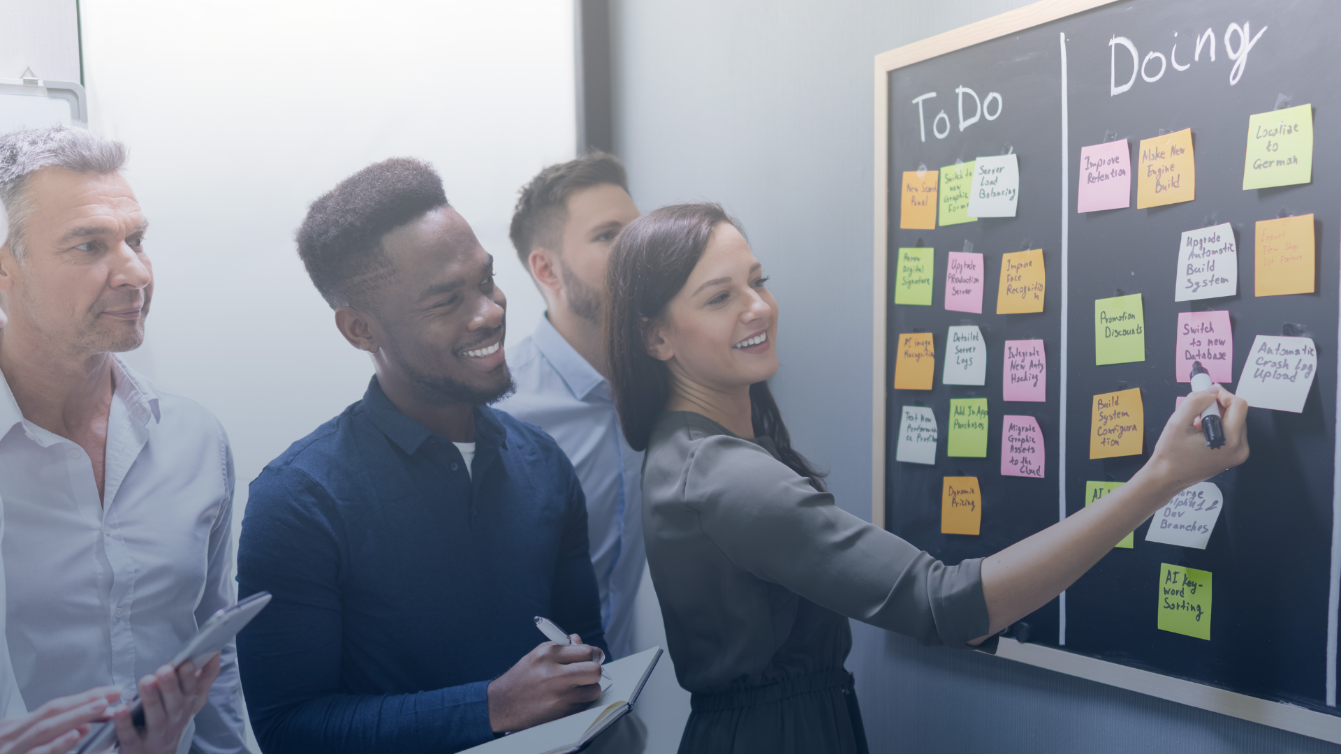 Women standing at agile board holding sticky note while three men look on