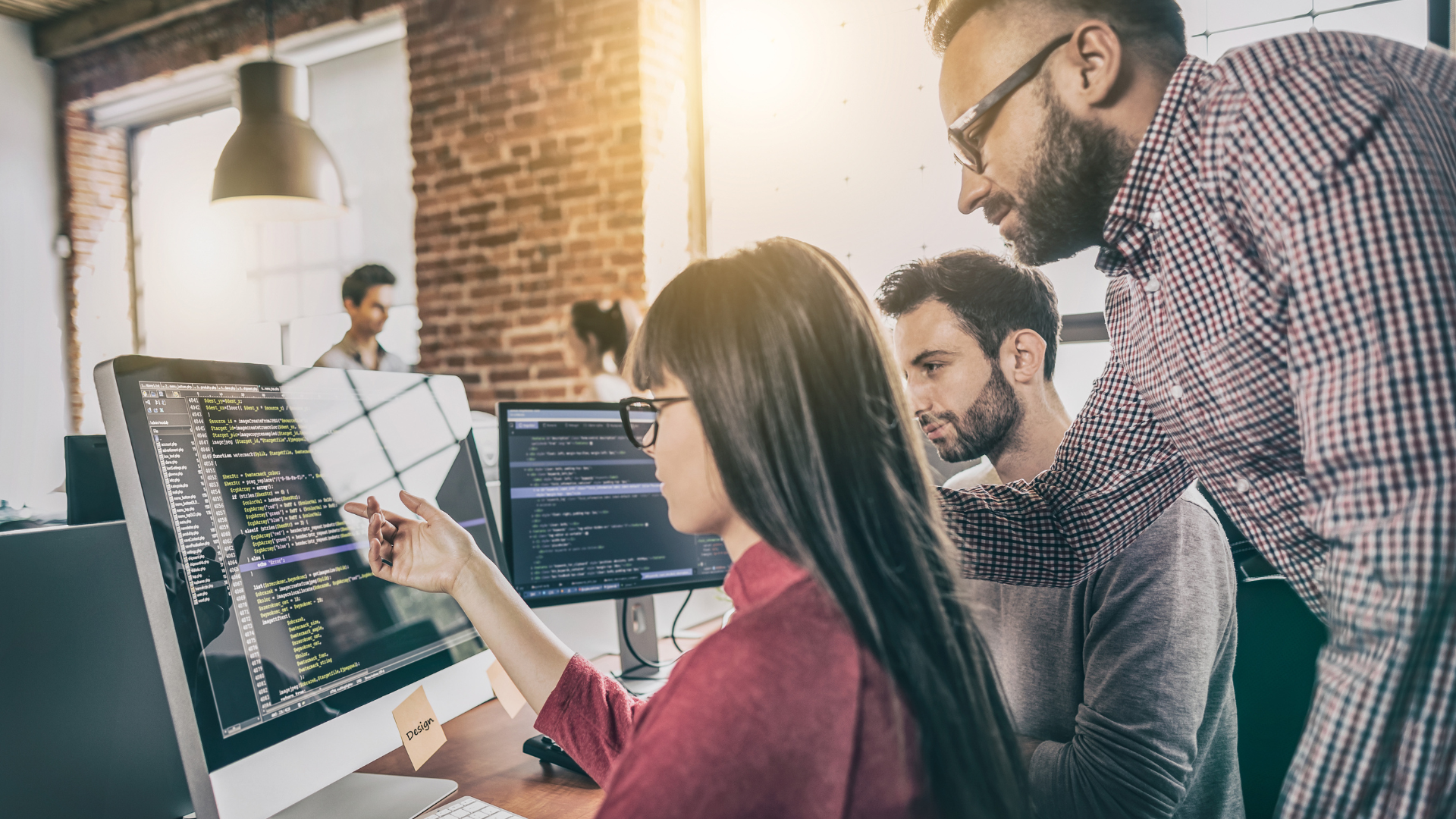 Woman sitting at desk looking at computer monitor while two men look over her shoulder at same monitor